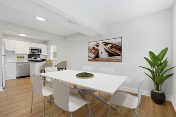 A dining room with a white table and chairs and a white kitchen with stainless steel appliances
